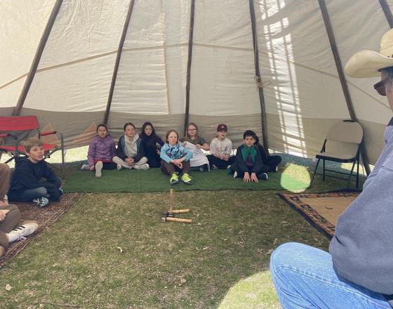 Children seated inside a large, round tent on a grassy floor.