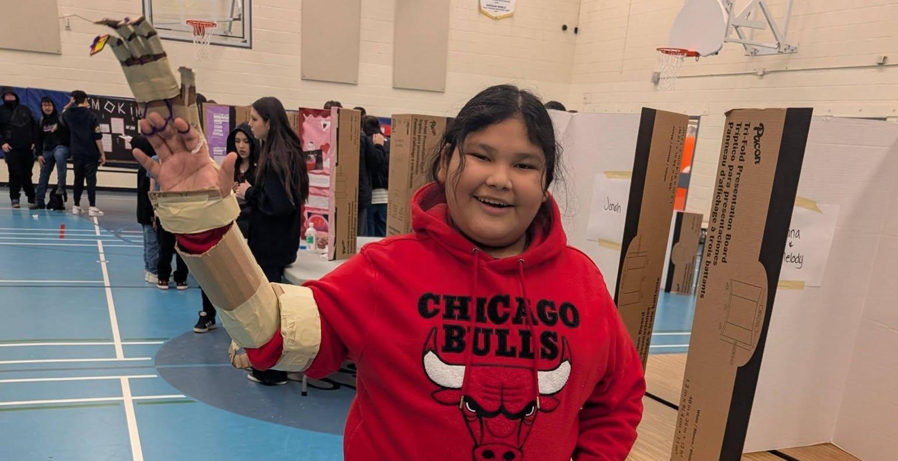A smiling student in a red Chicago Bulls hoodie stands in a gymnasium showing off a science project arm made of cardboard and tape.