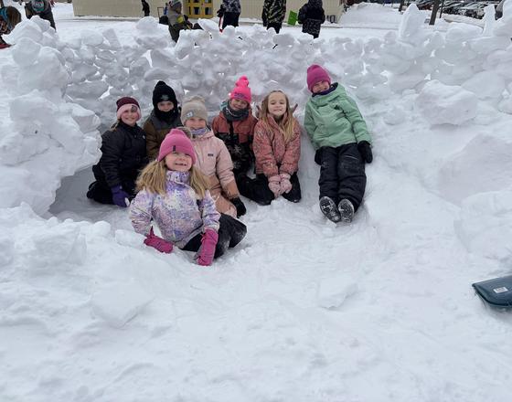 Group of children sitting in a snow fort, smiling and wearing winter clothing.