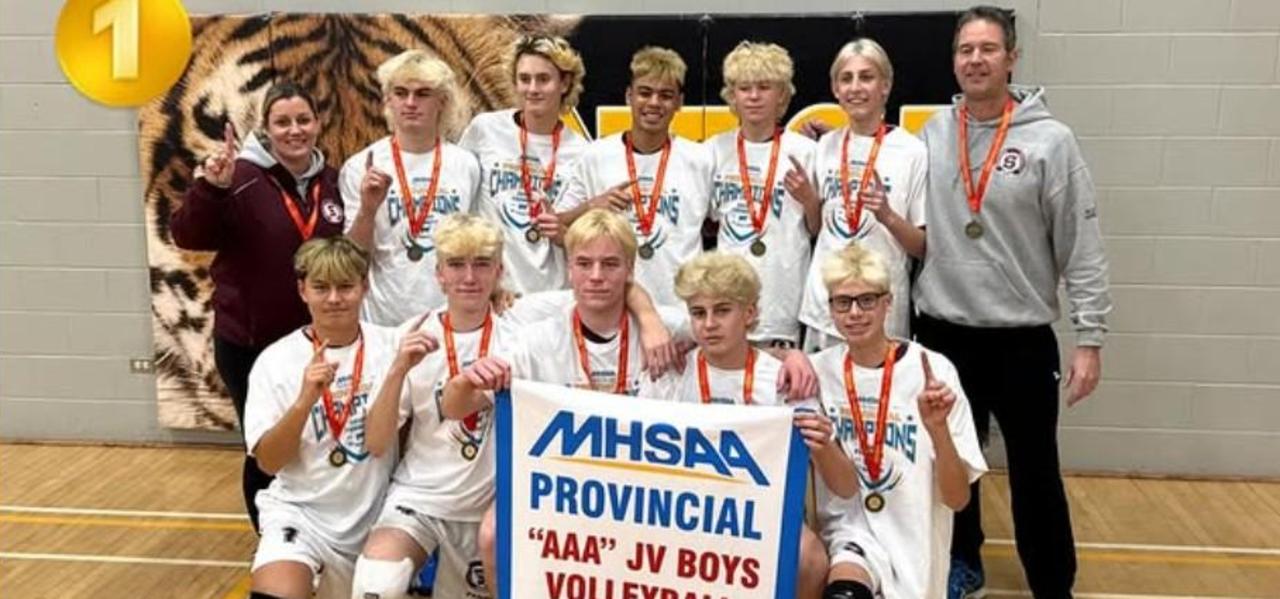 First row of 5 players kneeling on one knee holding a banner.  The banner reads MHSAA Provincial AAA JV Boys Volleyball Champions 2025-2026.  The second row has 5 players with a coach on each end.  Everyone has a medal hung around their neck.