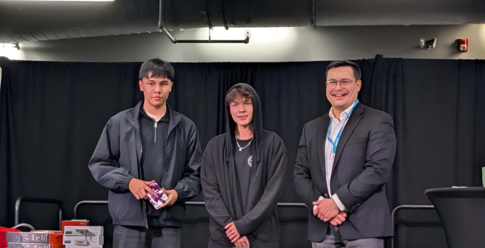 Two young men and a man in a suit standing behind a table with awards in front of a black curtain.