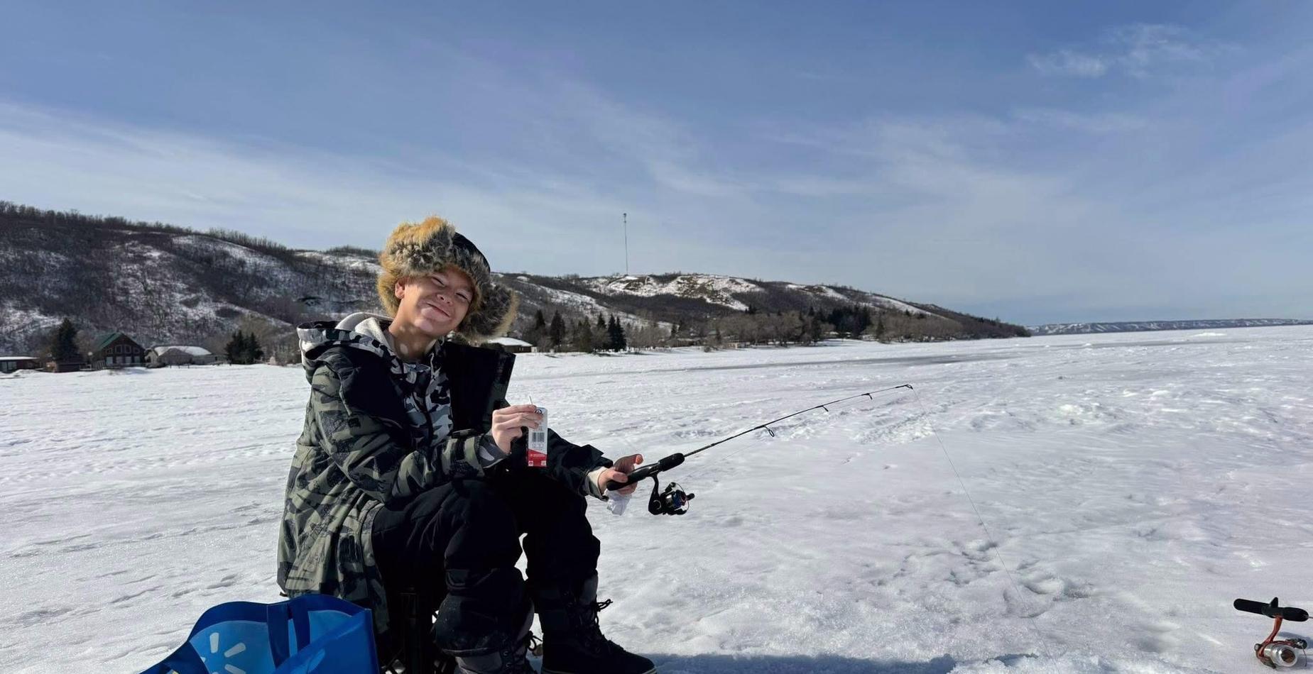 A person in winter gear ice fishing on a frozen lake under a clear blue sky.