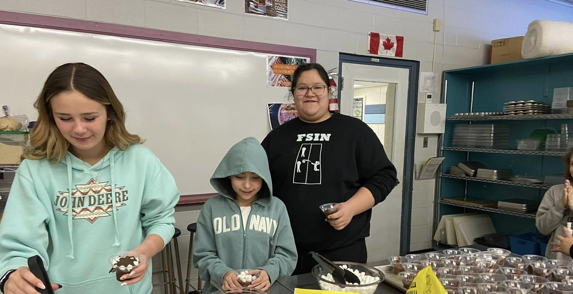 Three students in a kitchen workspace preparing cups of "dirt and worms" dessert using chocolate pudding, crushed cookies, and gummy worms.