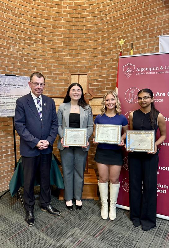 Three students standing with one adult in front of a ALCDSB banner and brick wall.