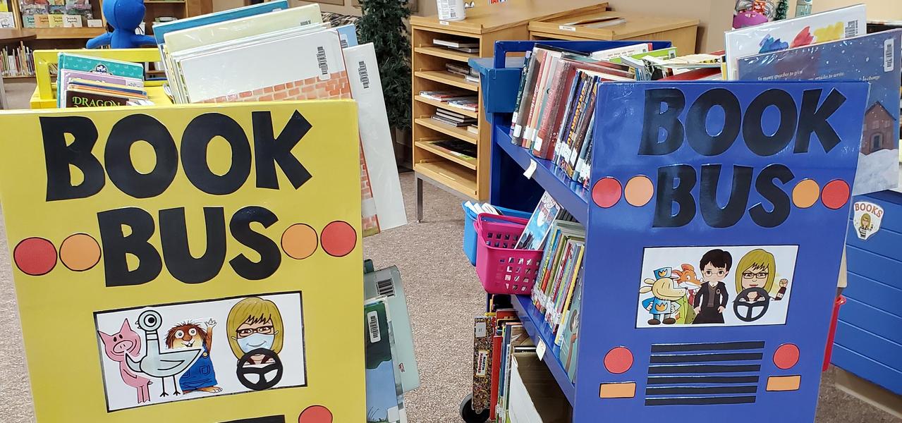 Two rolling carts, decorated as school buses, one blue and one yellow, titled Book Bus. Each cart was filled with books for students to read.