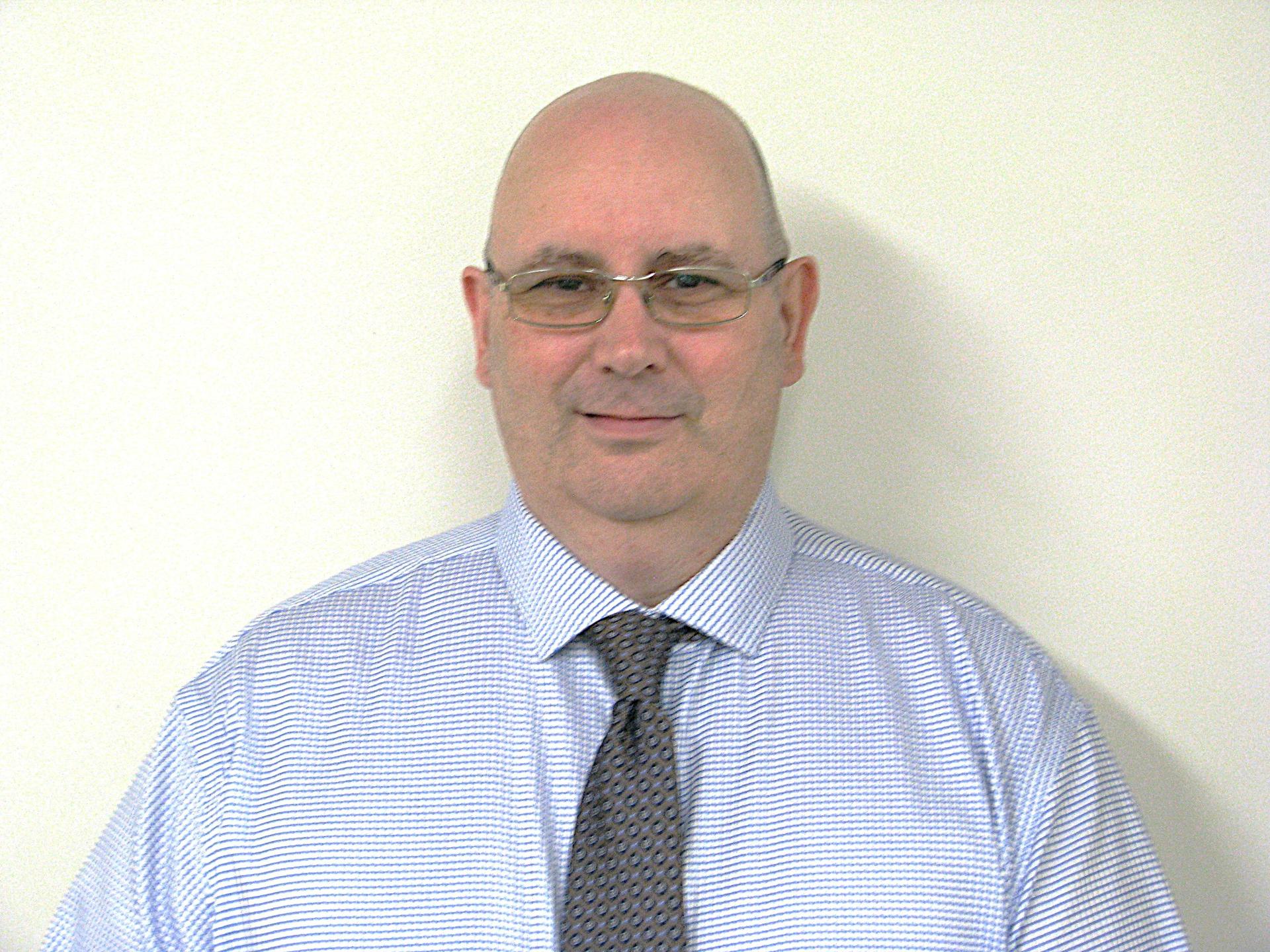 Head-and-shoulders portrait of a bald man wearing glasses, a blue striped dress shirt, and a patterned tie. He faces the camera with a slight smile against a plain light-coloured background.