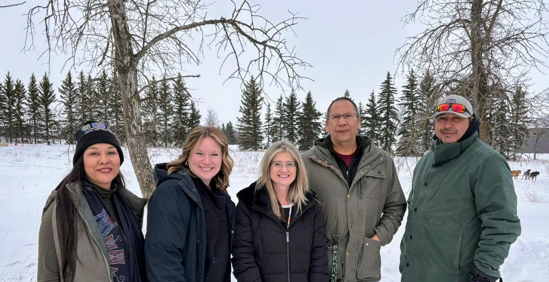 Five adults smiling together in a snowy outdoor setting with bare trees and evergreens.