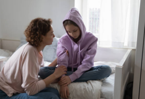 [Image: An adult sitting on a bed gently comforts a young person in a purple hoodie who looks down with a sad expression.]
