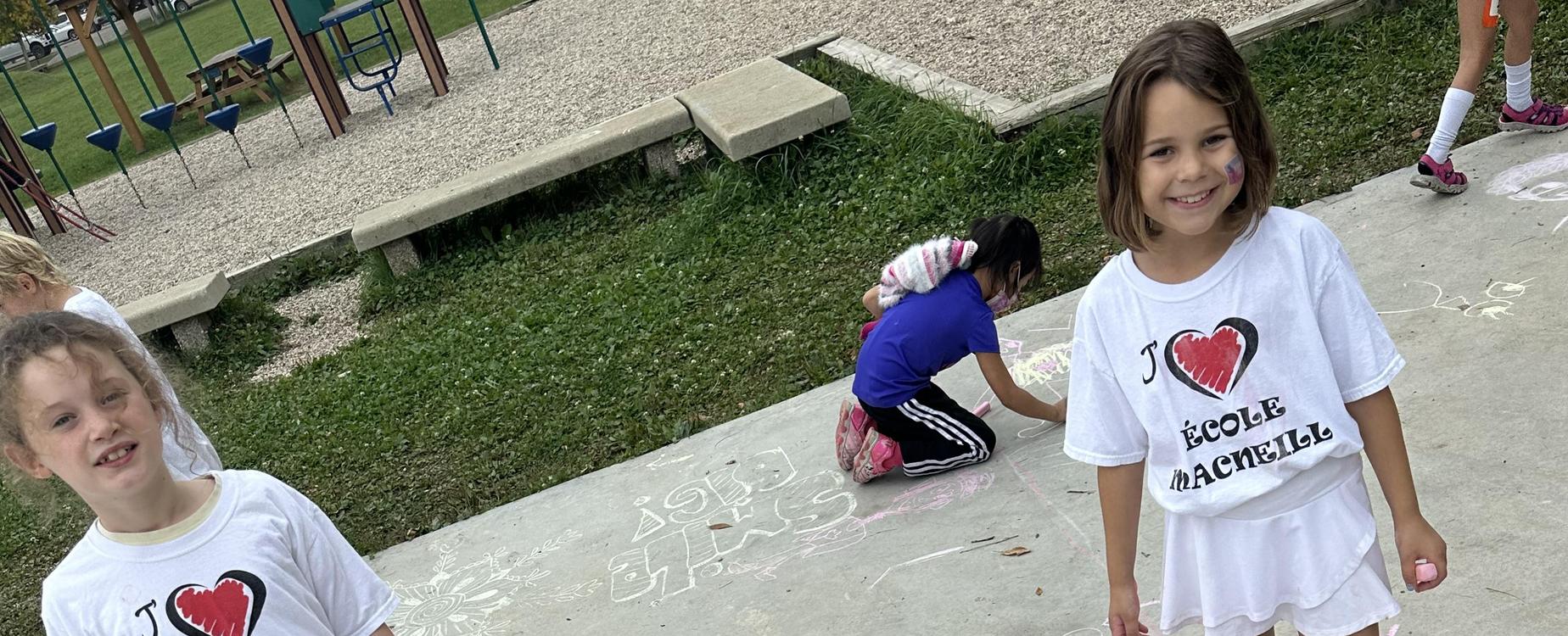 Two students with their J'aime École Macneill t-shirts. Showing off their chalk drawing of Terry Fox on the cement pad at school.