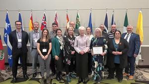 A group of fifteen adults stand in two rows in front of a display of Canadian provincial and territorial flags.