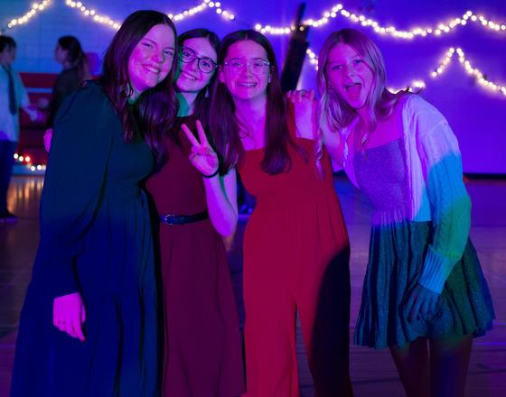 Four young women posing together at a dance, with colorful lights in the background.