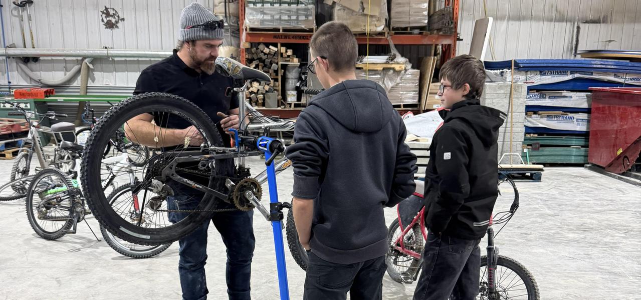 Two student standing, observing the middle years career development teacher instructing a bike repair.  They are in a large shop with cement floors.