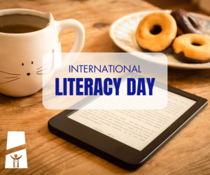A coffee mug and an e-reader on a wooden table. The text overlay reads “International Literacy Day.” Small white icons appear in the bottom corner.