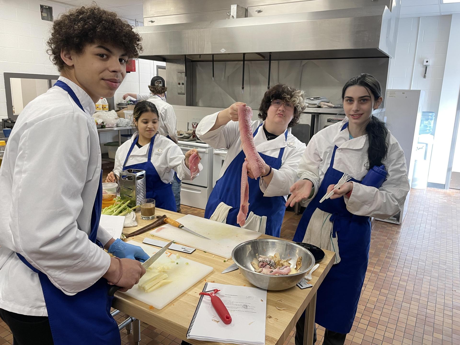 A group of people wearing white chef coats and blue aprons work together in a commercial kitchen. One person is slicing vegetables on a cutting board, while another holds up a long piece of raw meat over a metal mixing bowl. The table is covered with kitchen tools, a notebook, and ingredients. Stainless steel appliances and counters are visible in the background.