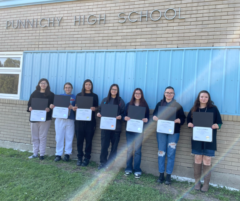 Seven Punnichy Community High School students stand in a row outside the school, each holding their Early Childhood Education Certificate of Achievement.