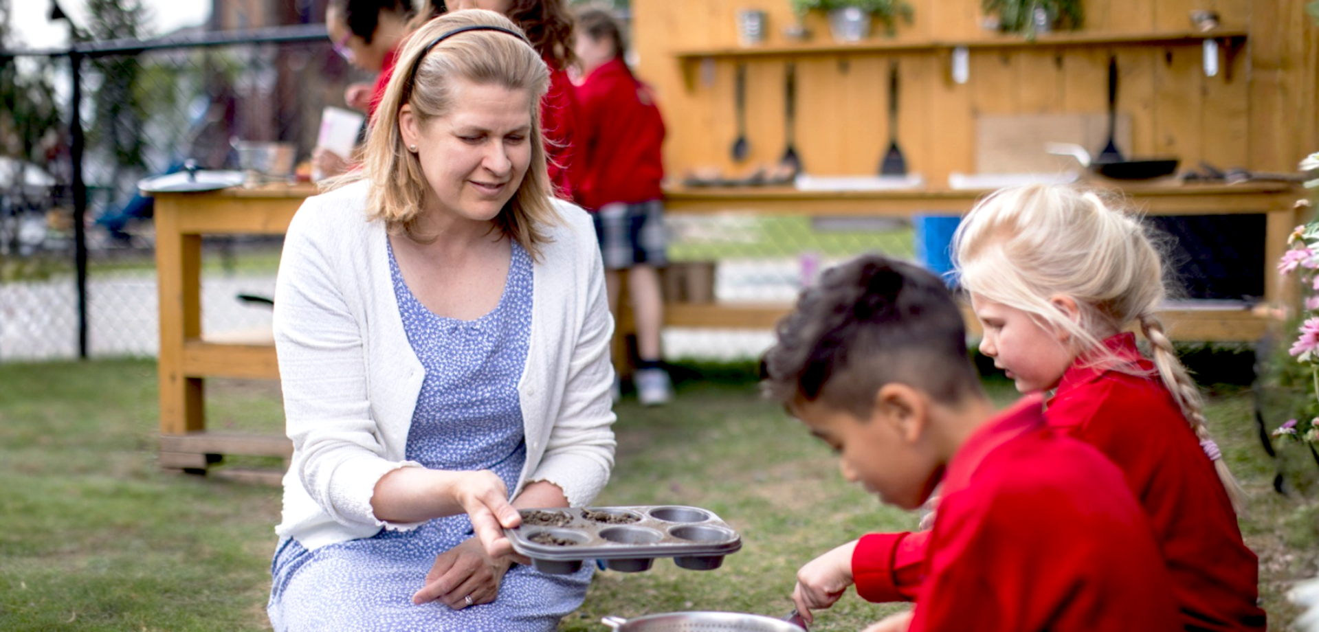 An adult woman interacts with two children during an outdoor cooking activity.