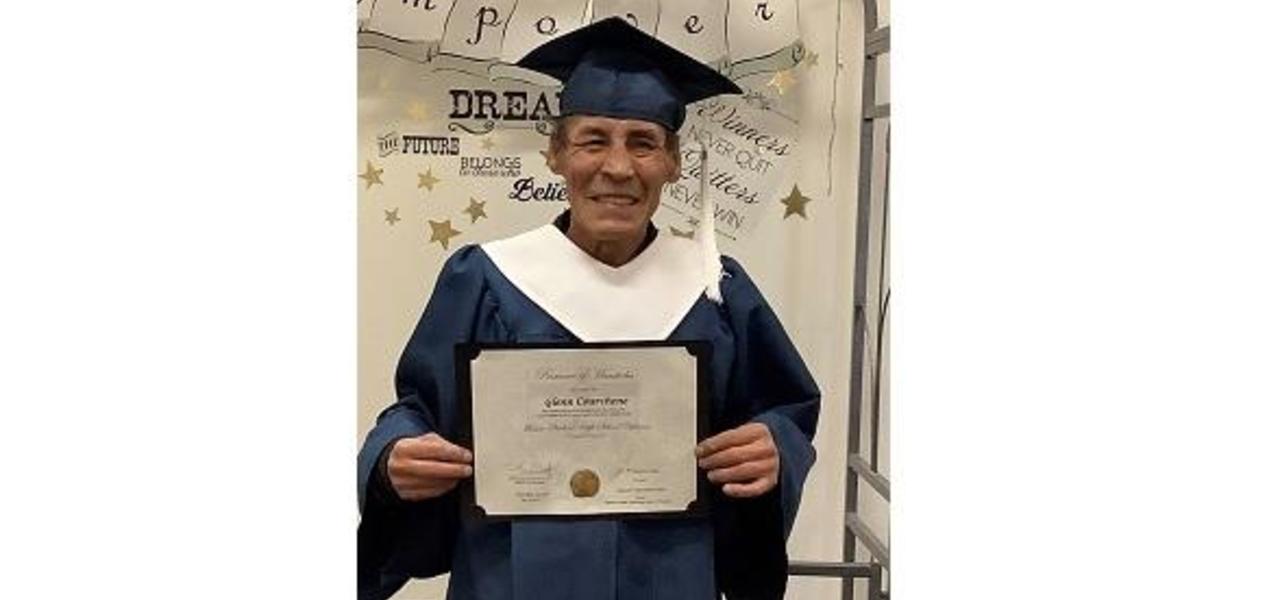 mature graduate holding grade 12 diploma. He is wearing a blue cap and gown.