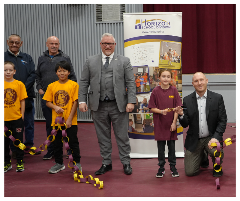 A group gathers inside the Kelvington School gym for the grand reopening ceremony. Two students stand at the front holding a long paper chain in school colours, joined by a student holding scissors for the ribbon-cutting. Behind them are community representatives, including MLA Chris Beaudry (centre in a grey suit) and a Horizon School Division banner displaying photos and the division map.