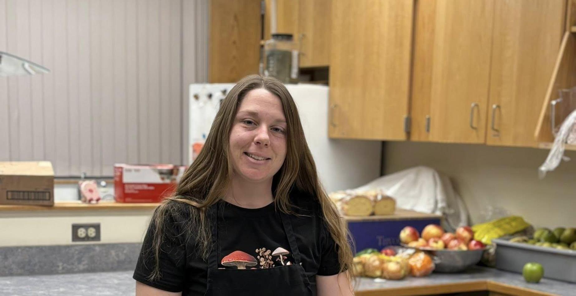 A woman in a black apron and gloves smiling in a commercial kitchen while placing unbaked croissants onto a parchment-lined baking sheet.