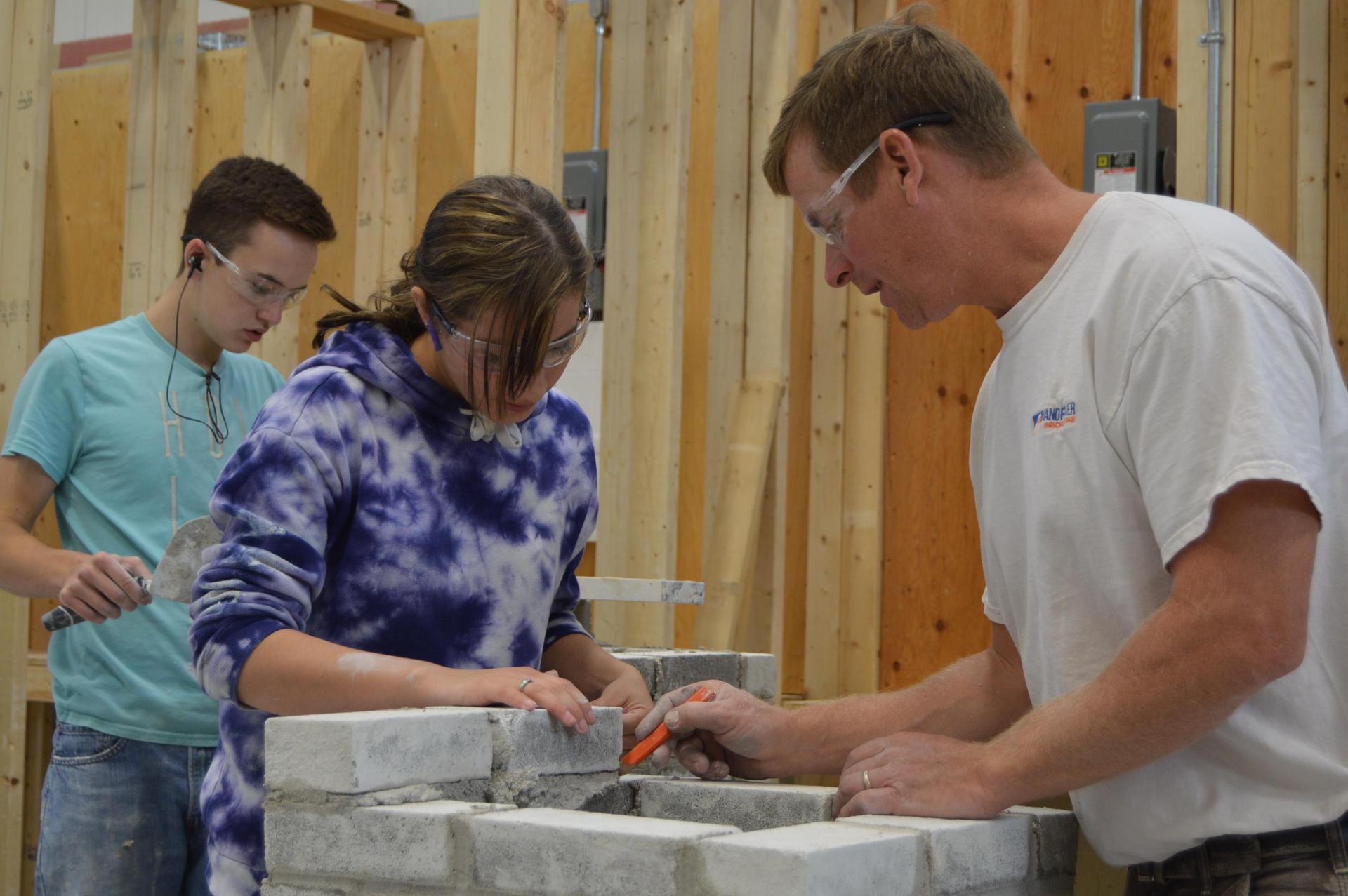 Two individuals are working on a partially built brick wall inside a workshop with wooden framing. One person is using a trowel to apply mortar between bricks, while another stands nearby holding a tool. A third person in the background appears to be mixing or handling mortar. Electrical boxes and wiring are visible on the wooden studs behind them.