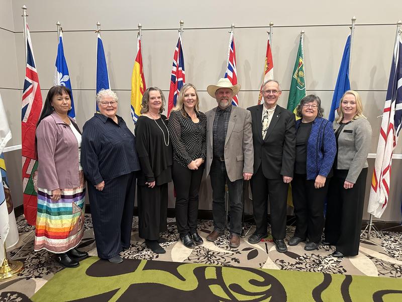 A group of nine adults standing in a row in front of a display of provincial and territorial flags. They are dressed in formal and semi-formal attire, with one man wearing a cowboy hat. The group is smiling and posed for a professional event photo.