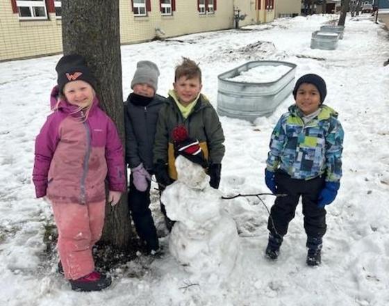 Four children smiling next to a snowman in a snowy outdoor setting.