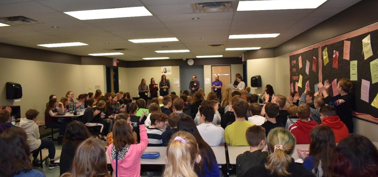 A large group of people including students and adults in a classroom. A group of adults at the front of the room presenting. Many students have their hand up to ask questions.
