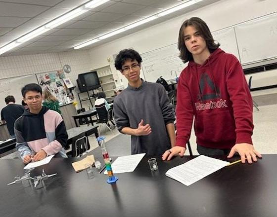 Three students in a science classroom with experiments on a table.