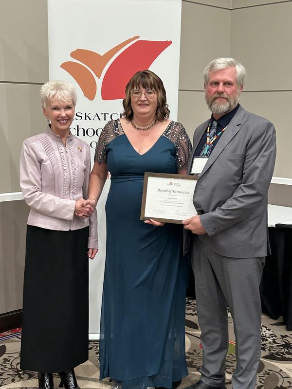 A woman holding an award certificate stands between two people—one shaking her hand—during a formal presentation. A Saskatchewan School Boards Association banner is displayed behind them.