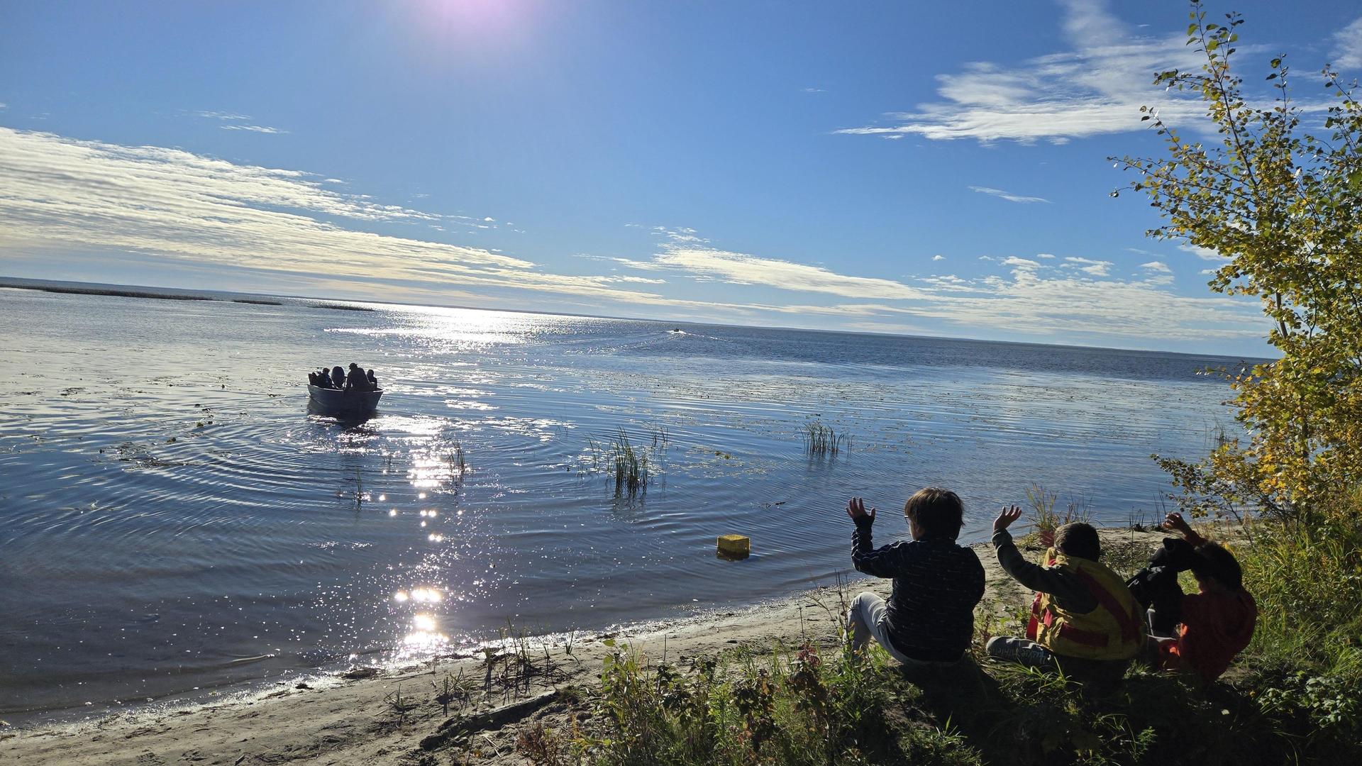 Three people waving at a boat on a shimmering lake.