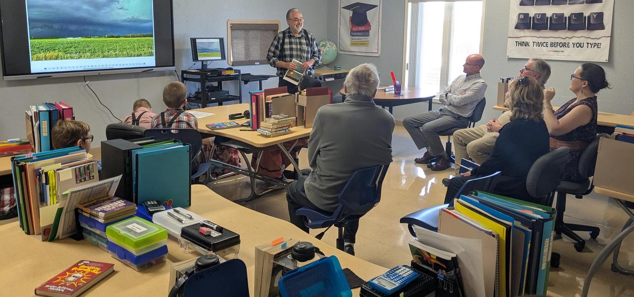 School Principal (gentleman with a beard) standing at the front of a classroom presenting to 5 adults and 3 children. There is a screen on at the front of the classroom.