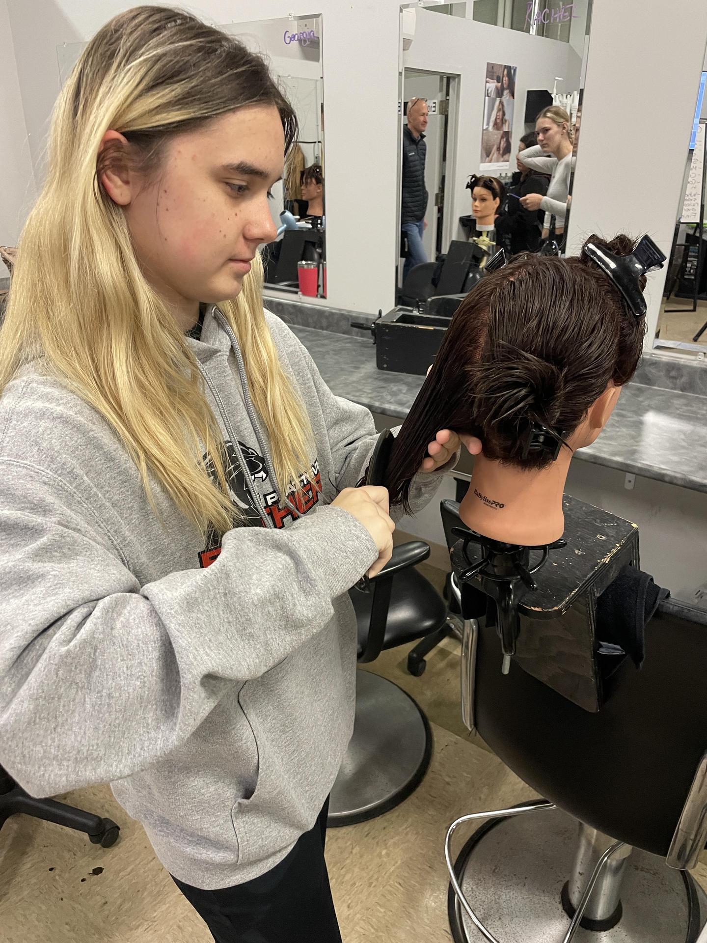 A person wearing a gray sweatshirt is styling a mannequin head with dark brown hair in a salon setting. The mannequin is secured on a stand, and the stylist is sectioning the hair with clips. Mirrors and salon chairs are visible in the background, along with other people working on similar tasks.