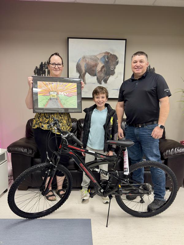 Bently stands behind his new bike, in between his teacher (holding his framed poster) and the Transportation Supervisor.