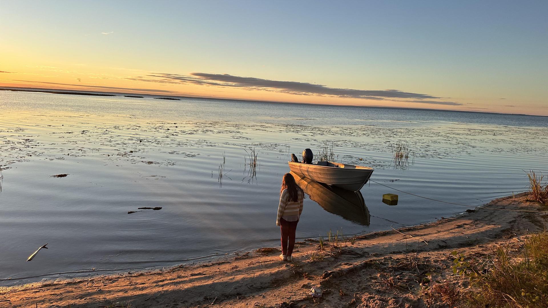 A person standing on the shore at sunset, with a boat anchored nearby.
