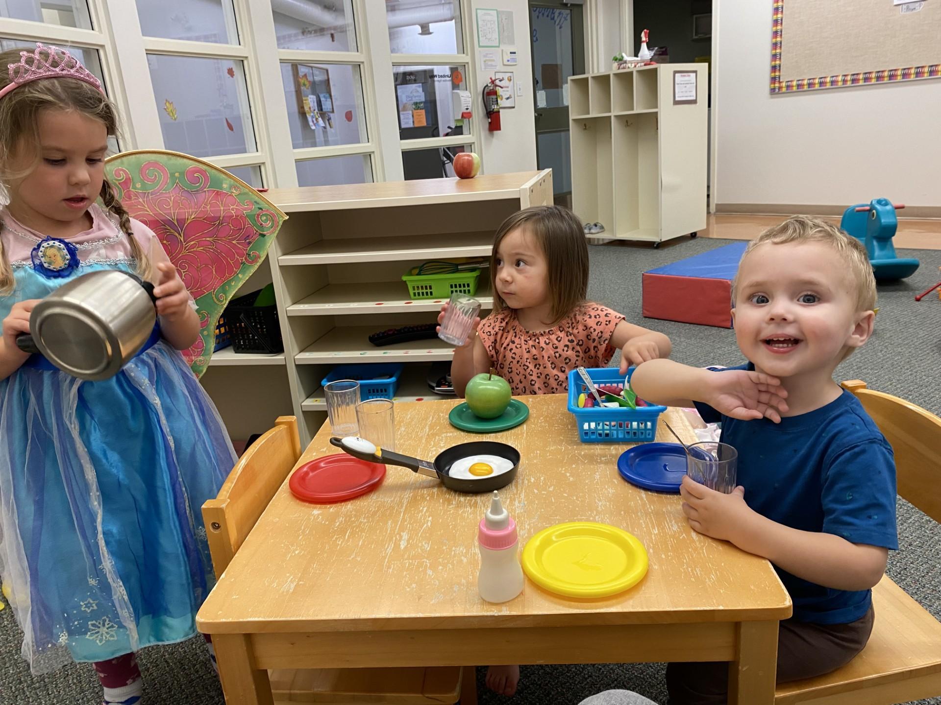 Three young children sit and play at a small table in a classroom, engaging in pretend play with dishes, cups, and toy food. One child wears a dress-up costume, while the others hold cups and smile, creating a playful and social learning environment.