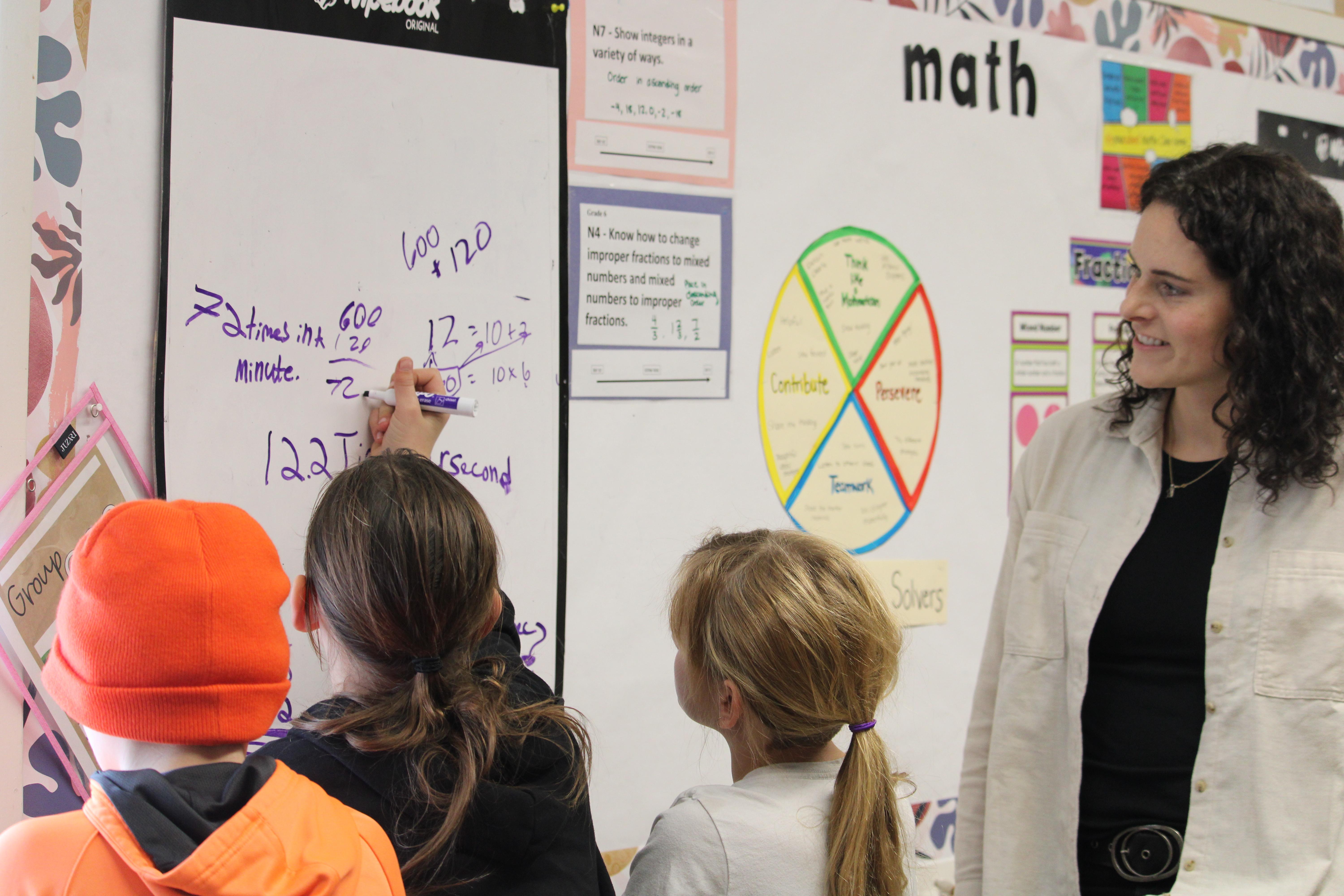 3 students work on a math problem while their teacher looks on.