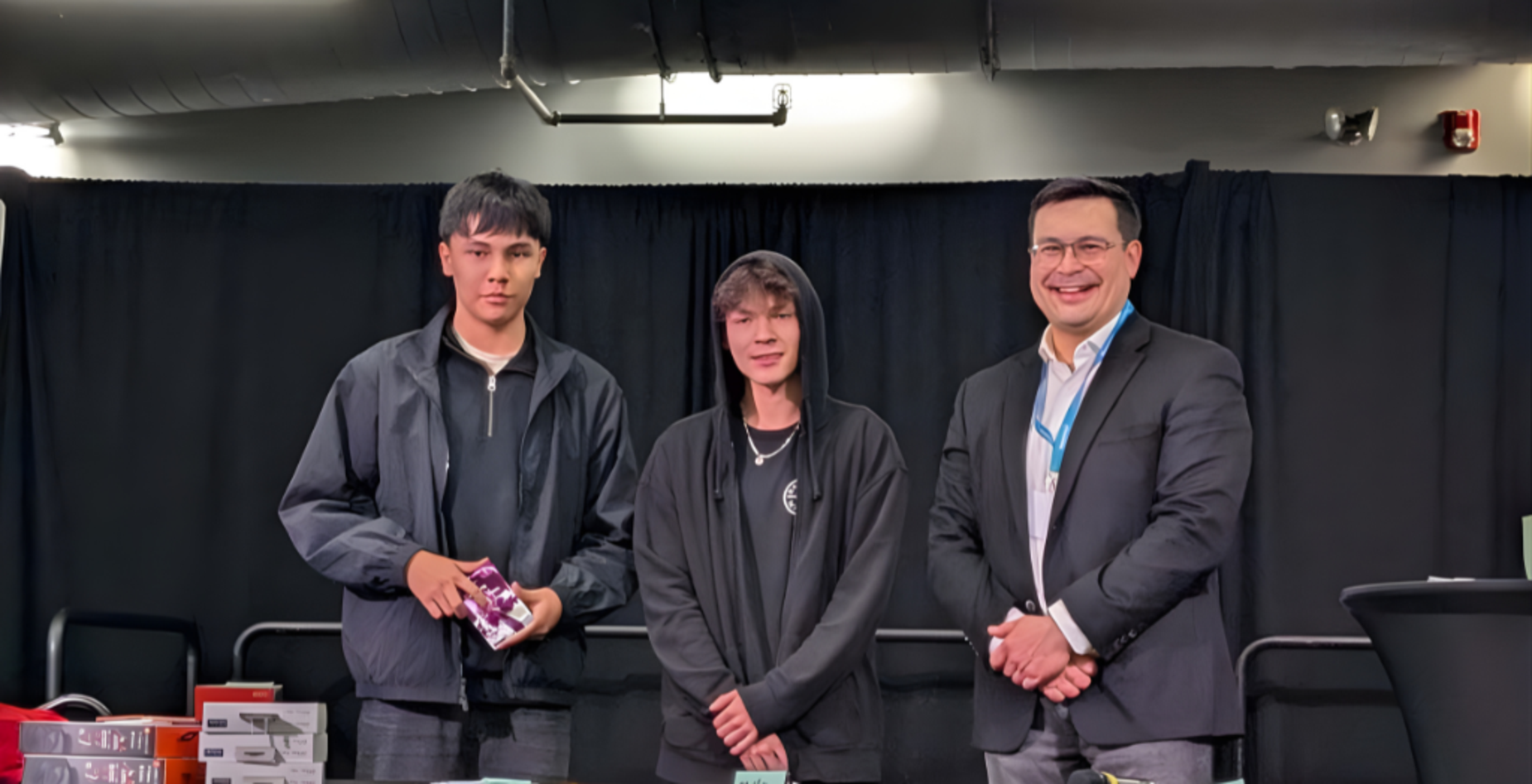 Two young men and a man in a suit standing behind a table with awards in front of a black curtain.