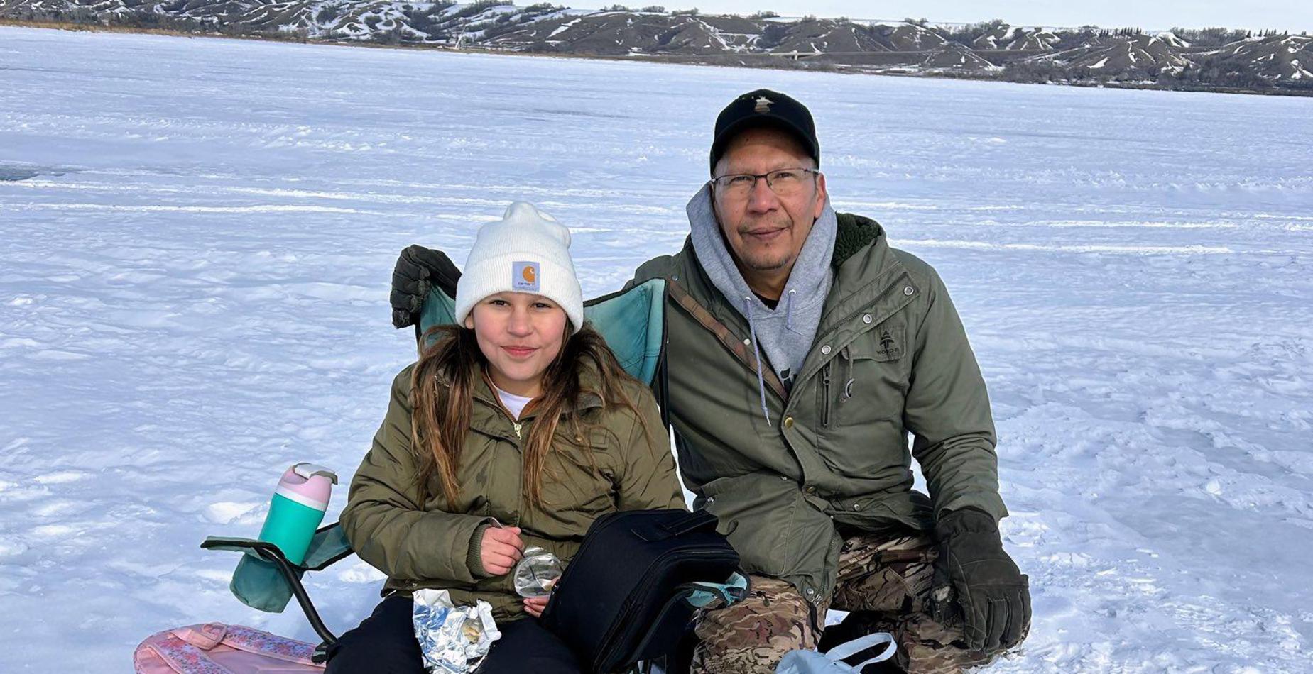 A child and an adult sitting together while ice fishing on a frozen lake.