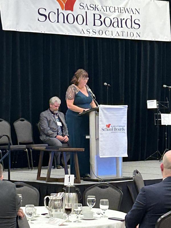 A woman in a teal evening gown speaks at a podium with a Saskatchewan School Boards Association banner. Seated behind her is a man in a suit, and audience members are visible in the foreground at banquet tables.