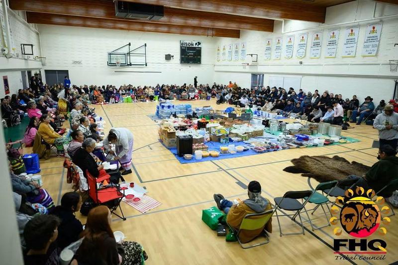 A wide-angle photo of a school gym showing students, elders, teachers, and community members seated around the gym walls and large tarps in the middle covered in various boxes and dishes of food for a celebratory feast