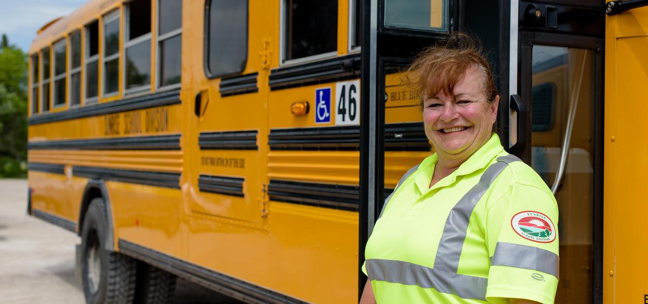 A female bus driver in a yellow high vis shirt standing in front of a school bus smiling.