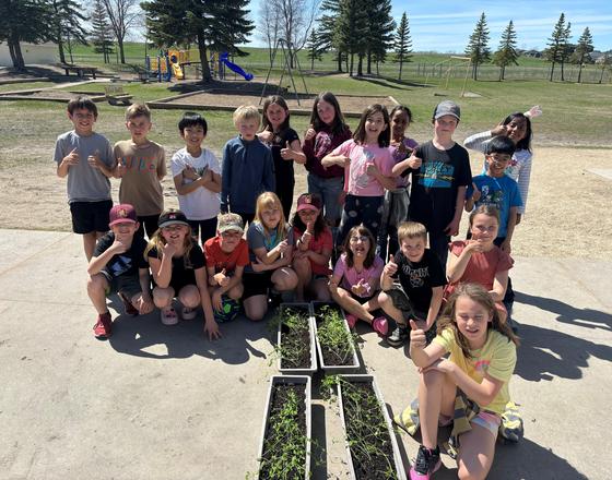Group of children outdoors, posing with thumbs up next to garden planters.