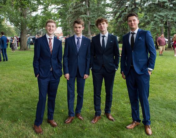 Four young men in suits standing on grass, smiling at the camera.
