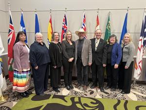 A group of nine adults standing in a row in front of a display of provincial and territorial flags. They are dressed in formal and semi-formal attire, with one man wearing a cowboy hat. The group is smiling and posed for a professional event photo.