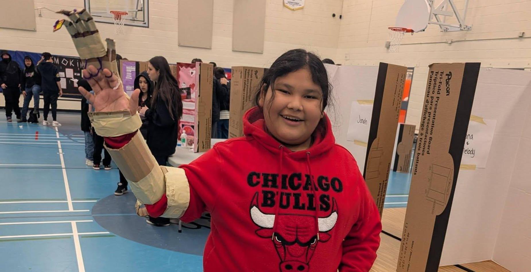 A smiling student in a red Chicago Bulls hoodie stands in a gymnasium showing off a science project arm made of cardboard and tape.