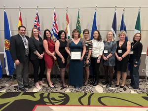 A group of eleven people standing in a row in front of a display of provincial and territorial flags. The woman in the centre is holding an award certificate, with colleagues and supporters standing on either side, all dressed in formal attire.
