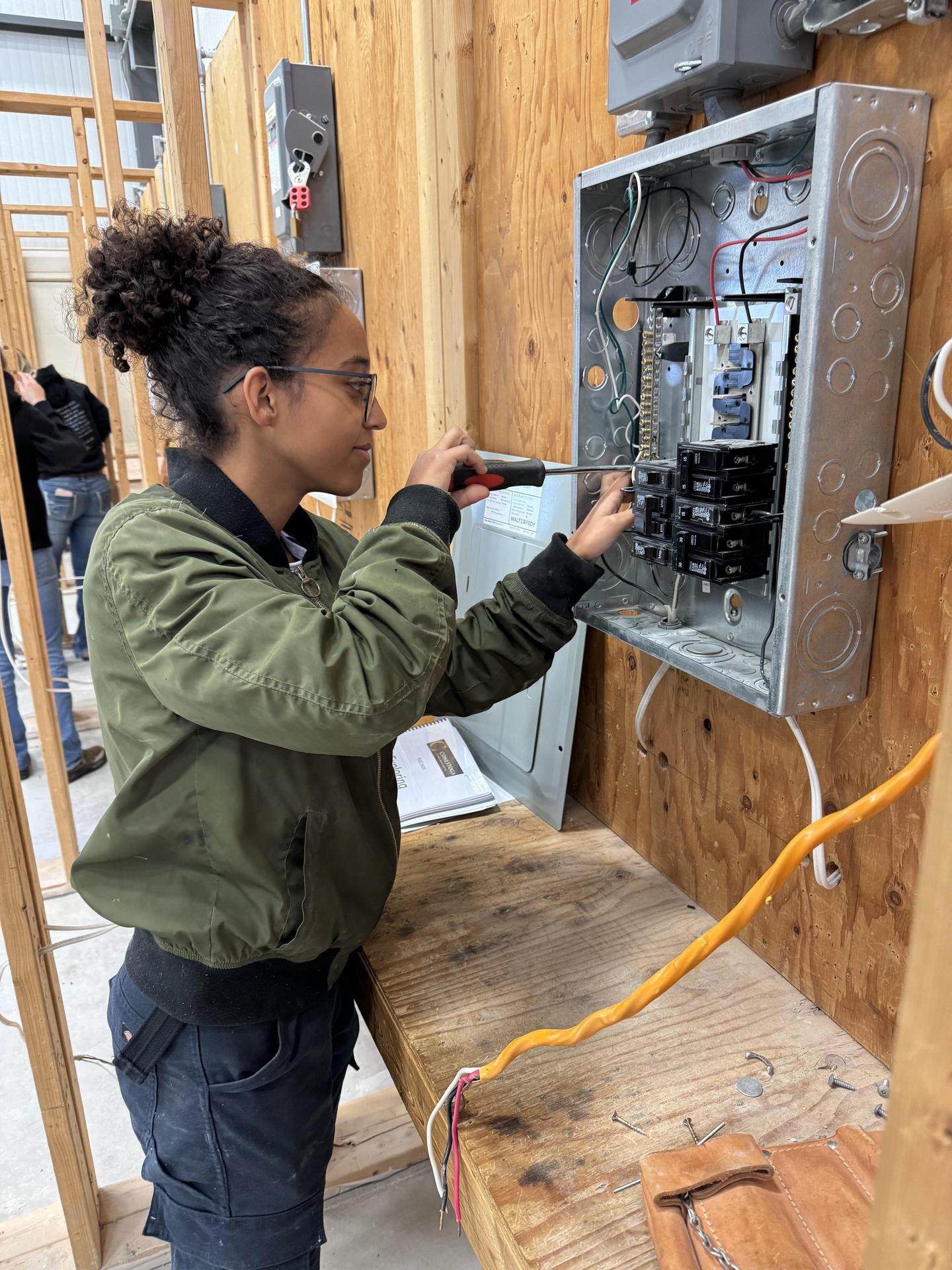 A person is working on an electrical panel mounted on a wooden wall inside a workshop. The panel is open, revealing wires and circuit breakers. The individual is using a screwdriver to adjust components inside the panel. An orange extension cord and a notebook are visible on the wooden workbench below.