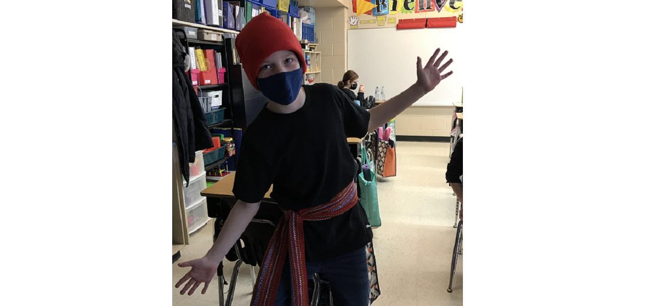 A student posing in a classroom wearing a red sache and toque, looking like a Voyageur.