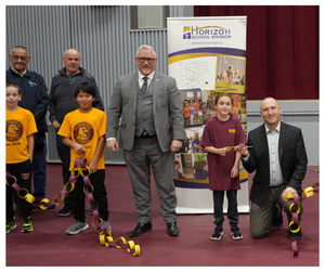 A group gathers inside the Kelvington School gym for the grand reopening ceremony. Two students stand at the front holding a long paper chain in school colours, joined by a student holding scissors for the ribbon-cutting. Behind them are community representatives, including MLA Chris Beaudry (centre in a grey suit) and a Horizon School Division banner displaying photos and the division map.