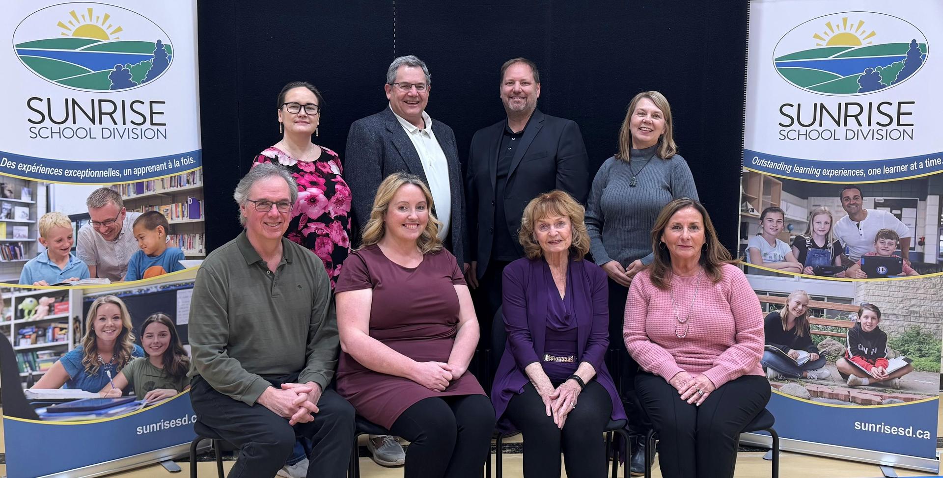 Four trustees sitting on chairs in the front row, 4 trustees standing behind them.  Sunrise Logo boards standing on each side of them with pictures of Sunrise students and teachers.